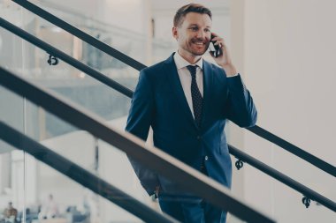 Zarif mavi takım elbiseli yakışıklı Avrupalı işadamı müşterisiyle cep telefonu görüşmesi yapıyor. Ofiste merdivenlerde dururken keyifli bir iş görüşmesi yapıyor. Akıllı telefondan konuşuyor.