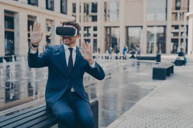 Sanal gerçeklikteki projeleri gözden geçiren, kürsüde otururken taşınabilir VR cihaz gözlüğü kullanan, el hareketleriyle hareket eden ve gülümseyen, simülasyon uygulamasını test eden, resmi takım elbiseli neşeli genç işletme sahibi.