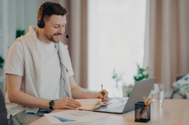 Kulaklık takmış gülümseyen bir adamın internet üzerinden video dersi alması, evde kazanç sağlarken deftere bazı notlar yazması. Halk ve uzak eğitim