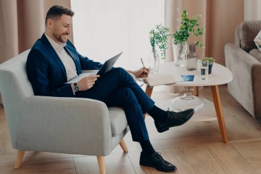 Side view of smiling bearded businessman in suit watching online business video while sitting in armchair and working remotely, using laptop and studying online. Business people lifestyle concept