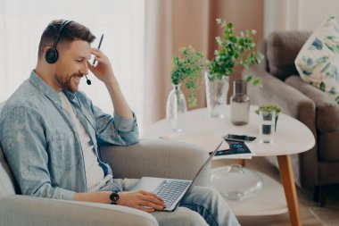 Side view of happy bearded freelancer in headphones sitting in armchair in living room of his apartment and working on laptop while having online meeting. Freelance and distance education concept