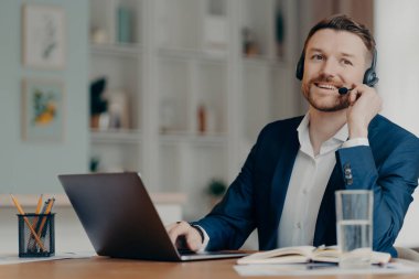 Happy male entrepreneur in suit working from home, successful businessman in headset talking with colleagues online through web conference while sitting at his cozy workplace. Remote work at home