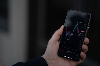 İnternet üzerinden ticaret için mobil uygulama. Ekranda mali ön plan grafiği olan akıllı telefon tutan erkek, borsa verilerini kontrol eden tüccar ya da yatırımcı, açık havada dururken, seçici odaklı