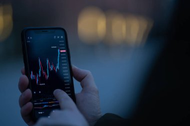 Erkek el, açık havada dururken gerçek zamanlı ön x grafikli akıllı telefon ekranına dokunuyor. Finansal grafiği olan cep telefonuna odaklanıyor. Mobil uygulamada borsa verilerini kontrol eden tüccar