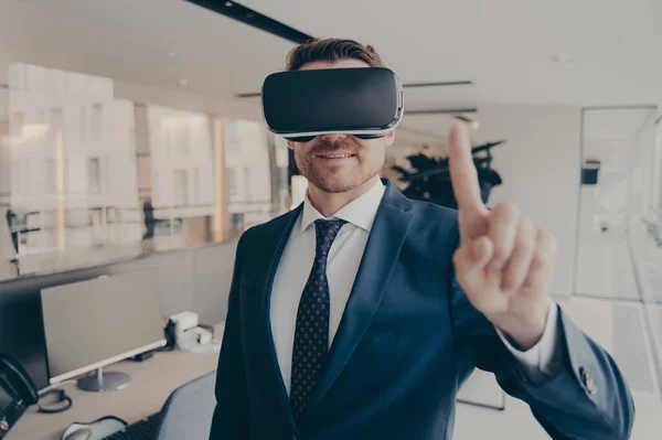 CEO man stands in office with VR headset and interacts with application virtual interface, pressing invisible buttons or selects menu items moving his forefinger in air. Cyberspace experience concept