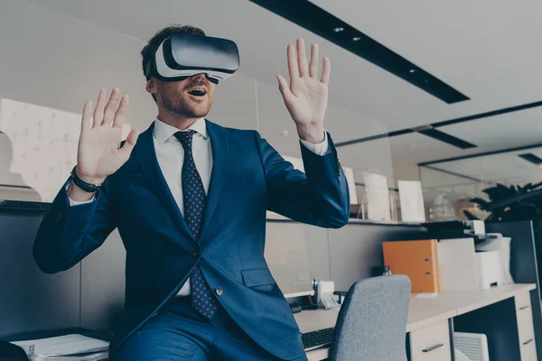 Amazed man in VR headset interacting with virtual objects or interface, sitting at edge of office table. Excited entrepreneur making 3D business presentation or meeting with virtual reality glasses