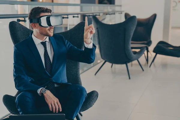 Young CEO sitting in office centre lobby with VR headset goggles , pointing with forefinger up in air, totally immersed in testing of virtual reality new business projects for company presentation