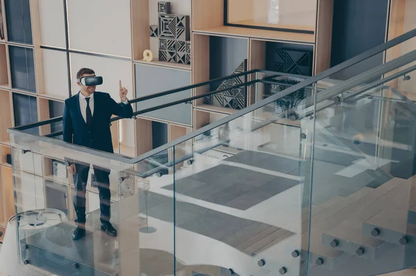 CEO director of gaming company testing prototype of VR headset glasses while walking around office center lobby and imitating movements in virtual reality, gesturing with index finger up in air