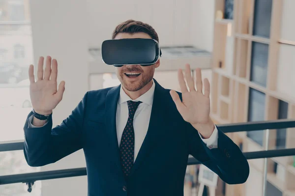 Smiling office worker in suit in 3d goggles enjoying cyberpace experience with raised arms, surprised excited businessman gesturing and touching object in virtual reality, using VR headset