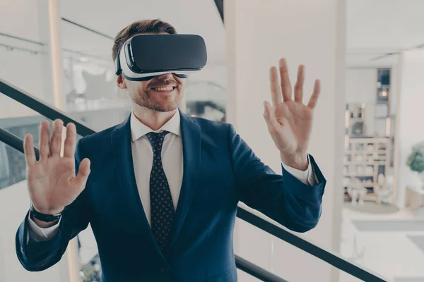 Young excited man office worker using virtual reality simulator at work, moving hands in air, being impressed of new innovative method in business, working with VR headset in digital actuality