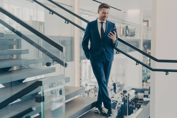 Stunning entrepreneur with neat beard in formal stylish suit standing on staircase in office lobby with smartphone in his hand, checking e-mails, financial reports or messages from business partners