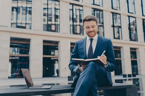 Professional smiling firm worker in formal suit writing down ideas with pen in note book while sitting on bench with his legs crossed, open laptop next to him, office buildings in blurred background