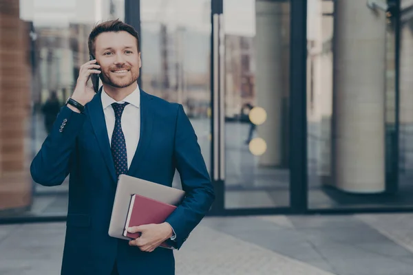 Happy male company worker leaving office building holding notebook and laptop while talking on phone with his colleague, sharing good news about recent events, blurred background