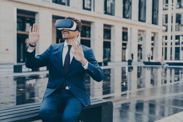 Man in formal outfit in VR goggles, trying to reach something in virtual reality with his hands in front of him, sitting on bench in front of fountain and office buildings outdoors during coffee break