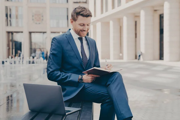 Successful business owner in formal wear writing down information he is receiving from conference call on laptop, sitting on bench with crossed legs and working remotely, doing freelance job outdoors