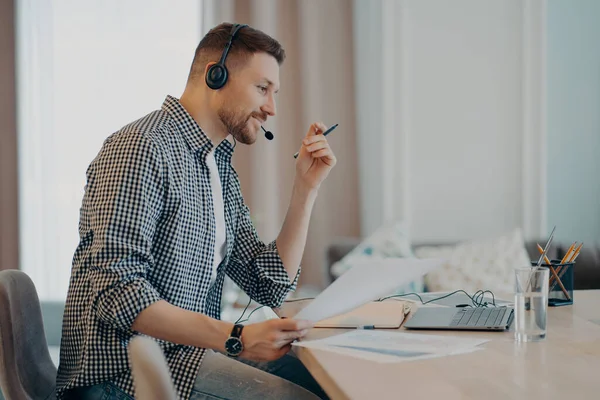 Side view of happy guy worker in casual clothes and headset having video call on laptop while working remotely during quarantine, male freelancer talking at online meeting. Distance work concept