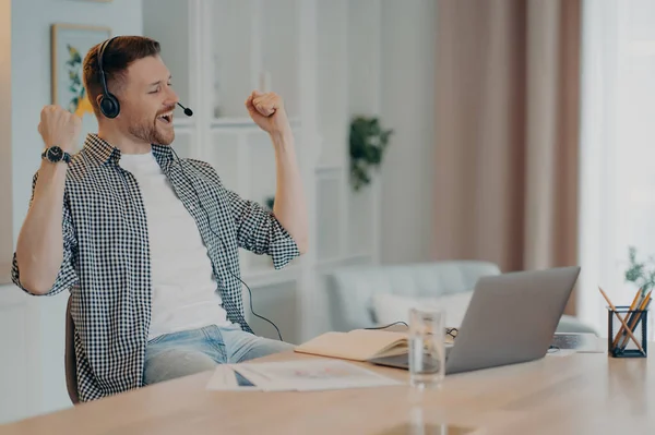 Happy successful man clenches fists celebrates triumph works online wears headset poses at desktop with laptop computer dressed casually. Overjoyed male student listens educational online lecture