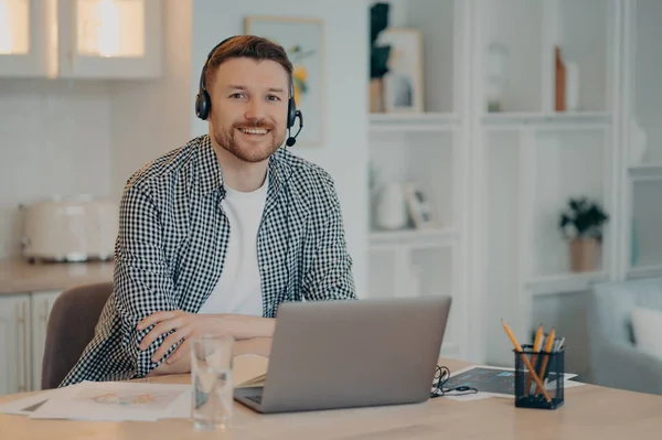 Horizontal shot of bearded man sits at desktop wears headset sits at desktop uses headphones studies or works from home prepared for video conference studies languages online poses at cozy interior