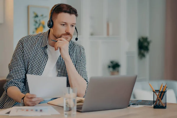 Serious man freelancer in casual wear holding document in hand and looking at laptop screen, analyzing information while working online in living room. Remote work and freelance concept