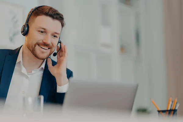 Smiling bearded businessman touching his hand on headset while having business meeting online, male freelancer using laptop computer at home. Freelance and distant work concept