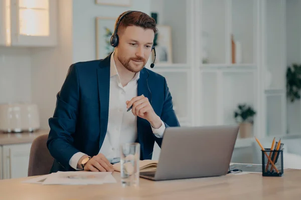 Cheerful male business consultant in headset working at home while talking with partner or client during online meeting or video call on laptop, sitting at his workplace. Online business