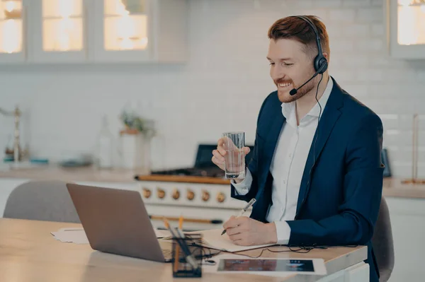 Smiling handsome businessman in suit sitting in headset having web conference on laptop while working at home, male freelancer with glass of water in hand looking at computer screen and smiling