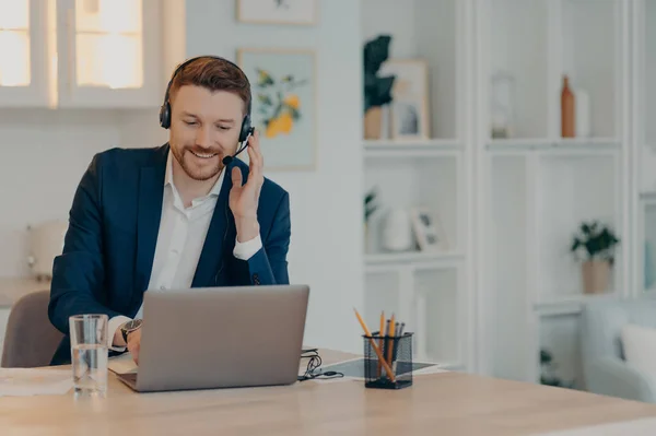 Happy male manager in suit working online on computer and using headset while sitting in living room and typing on keyboard, businessman working remotely from home. Distant work concept
