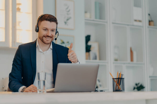Successful happy businessman in suit holding thumb up and looking at laptop screen with smile while communicating online with colleague, working remotely from home. Distant job concept