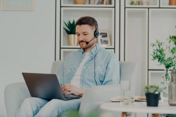 Contented guy in comfortable clothes using headset for video communication and holding laptop on his lap while staying home. Male freelancer working online. Distant work and freelance concept