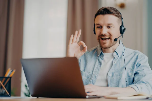 Happy bearded guy male freelancer showing ok sign gesturing during online call while sitting at his workplace with laptop in room, man working remotely at home. Freelance and online communication