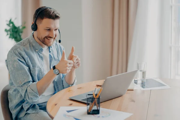 Joyful young guy looking at laptop screen and showing thumbs up while having online video call at home, looking at webcam and expressing positivity and agreement. Online communication concept
