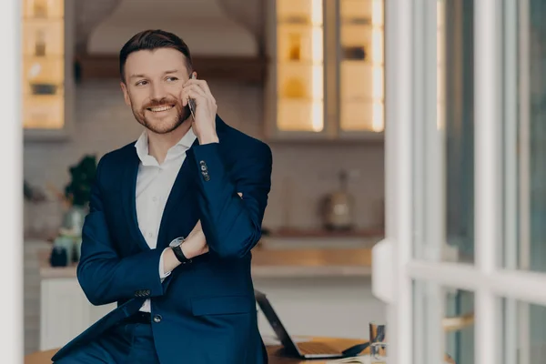 Corporate communication. Smiling professional bearded male entrepreneur has succssful business conversation keeps modern smartphone near ear poses near desktop dressed in formal suit focused away.