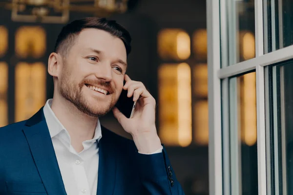 Mobile call. Handsome male executive manager with bristle has telephone conversation enjoys modern communication smiles happily looks thoughtfully into distance wers white shirt and elegant suit