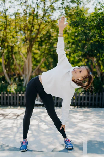 Vertical shot of glad sportswoman with combed pony tail leans aside keeps one arm raised up does exercises outdoors wears white hoodie leggings trainers relaxes on nature has active fitness training