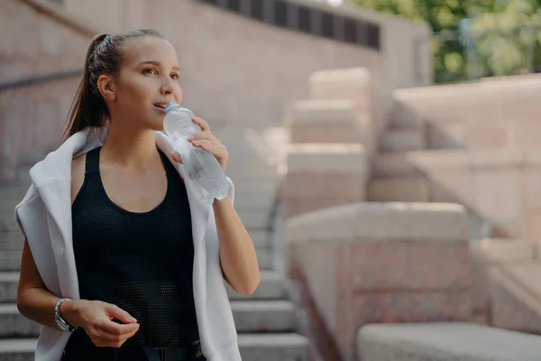 Keep water balance. Exhausted young sportswoman drinks cold refreshing drink after training takes break wears t shirt and white sweater around shoulders feels thirsty. Sport and health concept