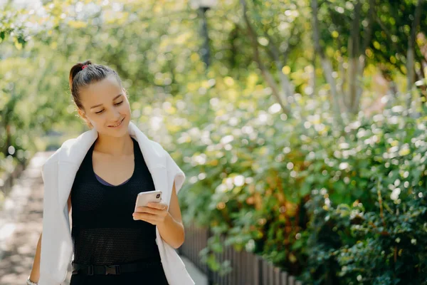 Pleased sporty woman with pony tail dressed in comfortable clothes focused at smartphone camera enjoys sport training outdoor walks in countryside checks message content. Lifestyle technology