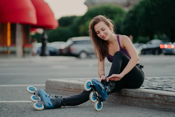 People leisure fitness sport recreation concept. Pleased young woman puts on rollerskates going to ride rolles in urban place has regular exercising goes in for dangerous sport adjustes laces