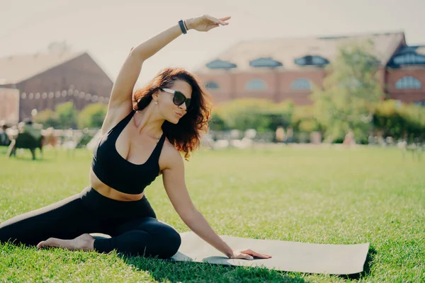 Image of motivated fit woman raises arm and does stretching exercises outdoors poses on fitness mat wears sunglasses and activewear trains actively. Fitness trainer goes in for sport outdoors