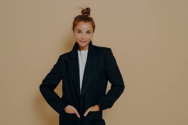 Siyah takım elbiseli, saçları yüksek topuz şeklinde bağlanmış, elleri ceketinin cebinde bej renkli arka planda poz veren çekici genç bir kadın. Moda tarzı stüdyo portresi. Zarif tarz kadınlar.