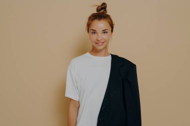 Beyaz tişörtlü, siyah ceketli, saçı topuz şeklinde bir omuz üzerine kıvrılmış, bej arka planda, kamerada gülümseyen boş bir arkaplan ile stüdyoda poz veren şık bir bayan öğrenci.