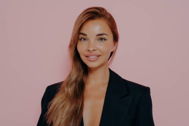 Çekici, gülümseyen, pozitif pozlarda duran, siyah şık bir takım giyen ve stüdyoda pembe arka planda kameraya bakan genç bir bayan. İş kadını ya da girişimci kadın konsepti