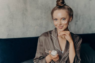 Kahverengi ipek pijamalı çekici bir kadın modelin sabah uyandıktan sonra elinde krema kabıyla yatağında otururken ve kozmetik ürünler uygularken yakın plan fotoğrafı.