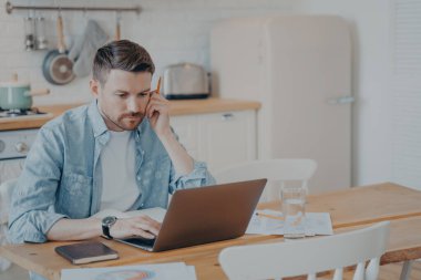 Ciddi şekilde odaklanmış genç sakallı iş adamı ya da serbest çalışan mutfak masasında oturuyor ve dizüstü bilgisayarda yazıyor, online proje üzerinde çalışıyor, araştırıyor ya da müşteriye e-posta yazıyor.