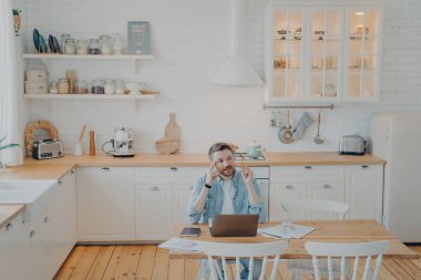Genç ofis çalışanı bilgisayarla uzaktan çalışıyor, mutfak masasında oturuyor, önemli bir projeyle görevlendiriliyor, çözümü bulmaya çalışıyor, not alıyor ve not alıyor.