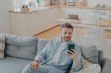 Evdeki rahat koltukta dinlenirken akıllı telefon kullanan beyaz bir genç, oturma odasında kanepede oturan yakışıklı bir adam, internette geziniyor ya da arkadaşlarıyla mesajlaşıyor.