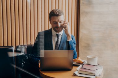 Mavi takım elbiseli gülümseyen genç bir işadamı, laptopunda kahve içip kruvasan içerken P ile telefonda konuşurken. Serbest konsept