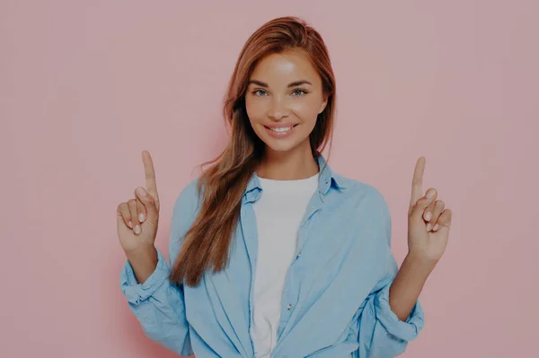 Happy young female points upwards with index fingers with broadly smile, shows awesome item on sale, wears white tshirt with blue shirt, isolated on pink studio wall. Promotion and advertisement