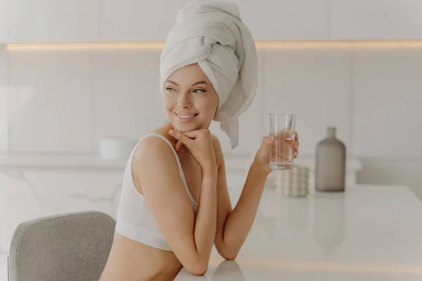 Side portrait of beautiful caucasian female sitting in kitchen after morning refreshing shower with head wrapped in bath towel, drinking mineral water and smiling. Healthy lifestyle concept