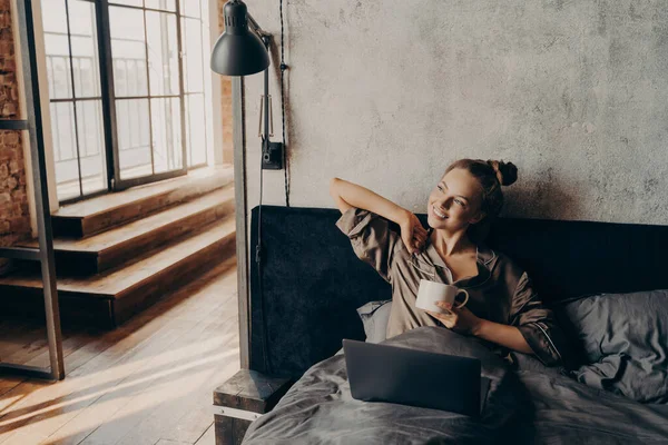 Happy positive young girl in satin pajama lying in bed at home after waking up from night sleep holding her morning cup of coffee and checking new emails on laptop or preparing to start remote work