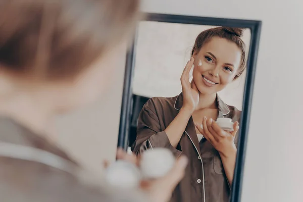 Reflection in mirror of beautiful woman in silk pajama smiling while applying face cream for deeper repair, protecting facial skin. Skincare, beauty and effective anti wrinkle treatment concept